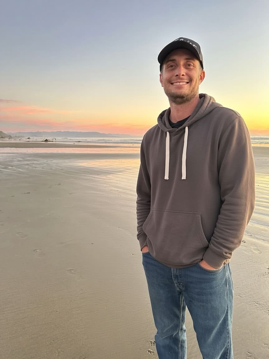 Garrett Doss (GRT) smiling on a beach at sunset in a hoodie and cap